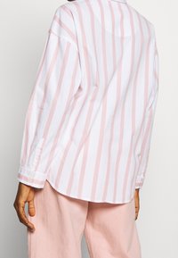 Long-sleeve shirt in white with pastel pink vertical stripes; relaxed fit, button detail on cuffs, soft cotton fabric, collar at neckline.