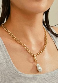 Collar de cadena de metal dorado con eslabones ovalados y un gran colgante de perla blanca irregular, llevado sobre una camiseta sin mangas de color gris claro.
