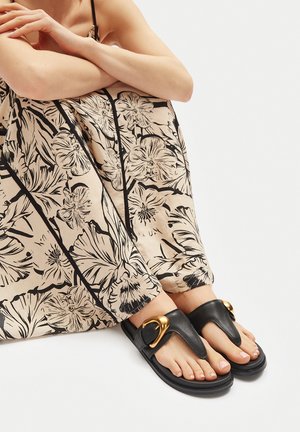 Persona con pantalones florales beige y sandalias de cuero negro con hebilla dorada, sentada con las piernas cruzadas contra un fondo blanco.