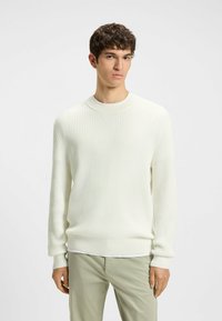 Maglione bianco a coste con scollo rotondo, caratterizzato da un tessuto testurizzato e maniche lunghe, indossato sopra una maglietta bianca e pantaloni verde chiaro.