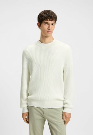 Weißer gerippter Strickpullover mit Rundhalsausschnitt, aus strukturiertem Stoff mit langen Ärmeln, getragen über einem weißen Unterhemd und hellgrünen Hosen.