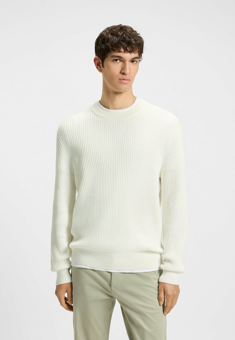Maglione bianco a coste con scollo rotondo, caratterizzato da un tessuto testurizzato e maniche lunghe, indossato sopra una maglietta bianca e pantaloni verde chiaro.