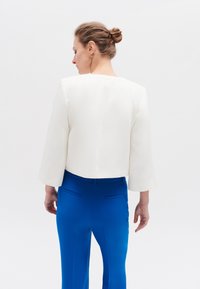 Witte cropped jas met brede mouwen, gecombineerd met blauwe op maat gemaakte broek met achterkantzakken. Gladde stofstructuur, minimalistisches ontwerp.