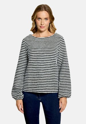 Femme aux cheveux blonds mi-longs porte un pull rayé noir et blanc avec de longues manches bouffantes et un jean bleu foncé.