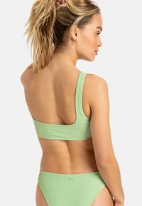 Conjunto de bikini de color verde claro con ribetes que incluye una parte superior de un solo hombro y una parte inferior de bikini a juego, mostrando una tela texturizada y acentos de hardware mínimos.