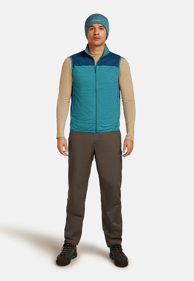 Muž v tyrkysovém beanie, tyrkysové zipové vestě, béžovém tričku s dlouhým rukávem, šedých kalhotách a černých turistických botách, stojící na jednobarevném pozadí.