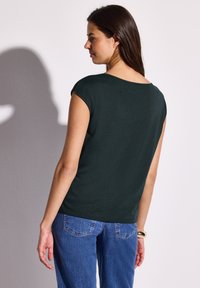 Tummanvihreä lyhythihainen paita, jossa on leveä kaula-aukko, sileä pinta ja rento malli, yhdistettynä sinisiin farkkuihin.