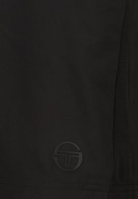 Indumento in tessuto nero con una texture liscia. Presenta un logo circolare nero opaco e dettagli su un pannello laterale. Design minimalista.