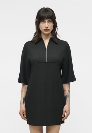 Γυναίκα με σκουρόχρωμα μαλλιά που φορά μαύρο φόρεμα με φερμουάρ μπροστά και μανίκια μέχρι τους αγκώνες, με ορατά τατουάζ σε κάθε αντιβράχιο.