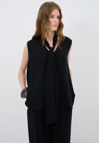 Haut sans manches noir avec un détail noué, présentant un col texturé et un tissu doux. Associé à un pantalon noir ample et un bracelet transparent.