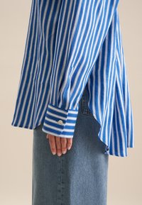 Blue and white striped shirt with long sleeves, buttoned cuffs, and a curved hem, paired with a denim skirt. Cotton fabric texture visible.