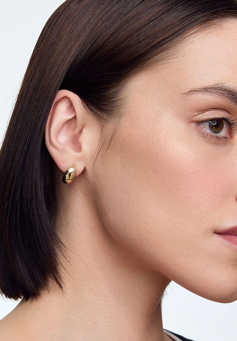 Złota kolczyk w kształcie okrągłym z gładkim, zaokrąglonym wzorem. Odbijająca światło powierzchnia z wypolerowanym wykończeniem, podkreślająca kształt ucha.