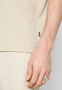 T-shirt beige à manches courtes en tissu texturé, avec une petite étiquette noire sur le côté. Une main avec une bague en argent repose sur le tissu.
