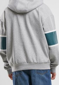 Sudadera con capucha gris de corte relajado, con detalles en teal y blanco en las mangas y un dobladillo acanalado. Material de algodón suave.