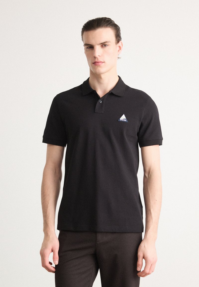 Juodas polo marškinėliai, pagaminti iš medvilnės, su klasikinėmis apykaklėmis, trumpomis rankovėmis ir mėlynu trikampio logotipu ant kairiojo krūtinės.
