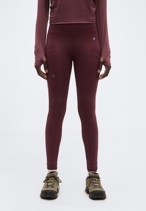 Άτομο που φοράει μπορντό κολάν με πλαϊνές τσέπες, ένα μακρυμάνικο τοπ σε αντίστοιχο χρώμα και ελιά πράσινα παπούτσια πεζοπορίας, στέκεται όρθιο.