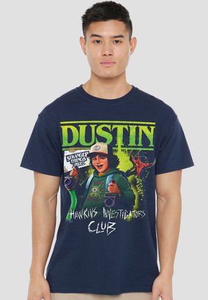 Young man wearing navy blue t-shirt with colorful graphic of Dustin from Stranger Things and text "Hawkins Investigators Club."