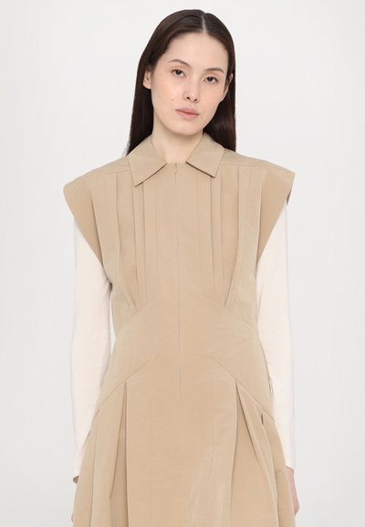 Rochie bej cu un design structurat, având un guler, pliseuri și cusături unghiulare. Purttă pe un top alb cu m�ânecă lungă. Textură netedă.