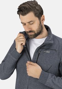 Graue Zip-Jacke aus einem weichen, strukturierten Stoff, mit hohem Kragen und einer Brusttasche mit Reißverschluss. Logo auf der Vorderseite angebracht.