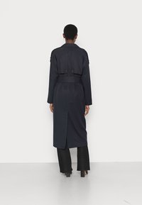 Marinblå trenchcoat med bälte i midjan, två knappar på ryggen och en slits i mitten bak. Slät tyg och långa ärmar.