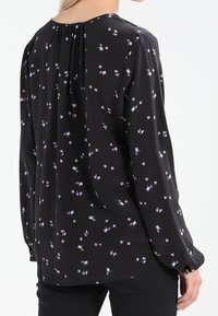Černá blůza s malým fialovým a bílým květinovým potiskem. Má sklady na zadní straně a dlouhé rukávy, vyrobena z měkké, lehké tkaniny.