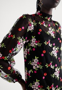 Vestido preto transparente com bordados florais em rosa, vermelho e verde. Mangas longas com punhos franzidos e gola alta. Design elegante.