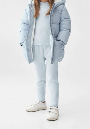 Barn som bär en ljusblå pufferjacka över en matchande ribbad tröja och byxor, med vita velcro-sneakers stående på ett vitt golv.