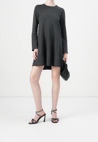 Femme portant une robe courte à manches longues de couleur sombre, des sandales à talons hauts noires à clous, et tenant un sac à main en cuir noir.