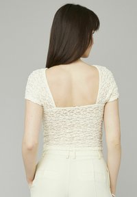 Un haut couleur crème avec un design en dentelle texturée, des manches courtes et un décolleté carré dans le dos, associé à un pantalon beige clair.