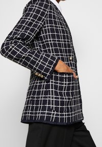 Marinblå och vit rutigt kavaj med texturerad tyg, två knappar och en ficka, med guldtonade detaljer på ärmsluten.