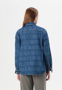 En kvinne med en blå denimjakke med et falmet tekstmønster på ryggen står mot en hvit bakgrunn. Hun har langt brunt hår.