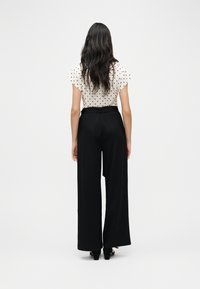 Γυναίκα με μακριά σκούρα μαλλιά φοράει λευκό μπλουζάκι με κοντά μανίκια και πουά μοτίβο, το οποίο έχει μπει μέσα σε ψηλόμεσο παντελόνι με φαρδιά μπατζάκια σε μαύρο χρώμα, στέκεται με την πλάτη προς τα εμπρός.