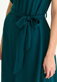 Täckande klänning i teal med ett mjukt, flödande tyg, som har ett uppknutet midjebälte. Designen inkluderar en slät yta och en avslappnad passform.