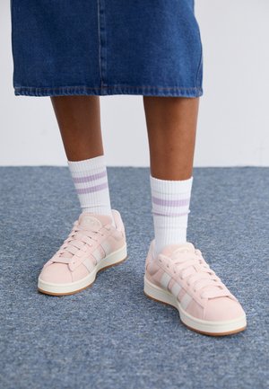 Jambes portant des chaussettes blanches à rayures violettes, baskets Adidas roses avec des rayures blanches, debout sur un tapis bleu à côté d'une jupe en jean.