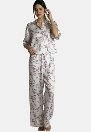 Femme portant un ensemble de pyjama blanc à fleurs, ample, avec des manches longues et un pantalon à jambes larges, debout pieds nus devant un fond uni.