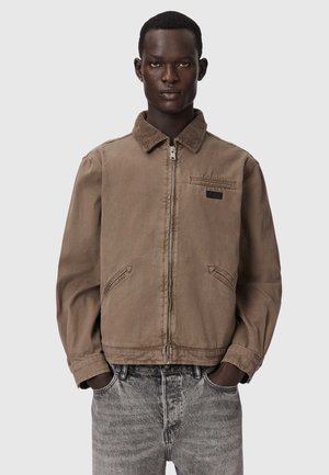 Mężczyzna w brązowej kurtce na zamek z kołnierzem i dwoma przednimi kieszeniami, w połączeniu z wyblakłymi szarymi dżinsami, ręce w kieszeniach, neutralne tło.