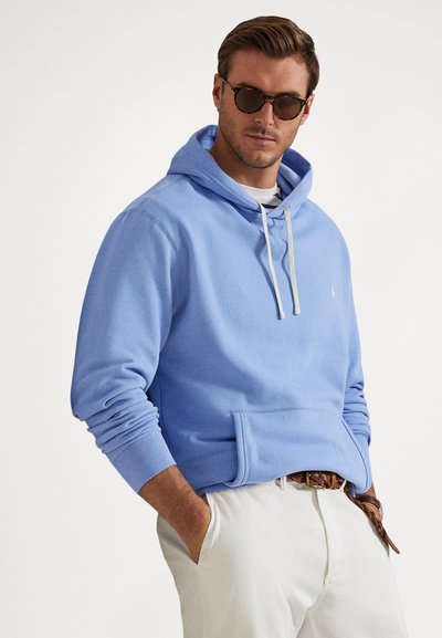 Šviesiai mėlynas džemperis su kišene priekyje, raišteliais ir ribinėmis rankogaliais, dėvimas ant baltos marškinėlės. Modelis dėvi šviesių spalvų kelnes.