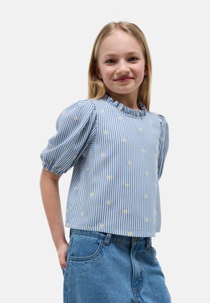 Fille souriante aux longs cheveux blonds portant un chemisier rayé bleu et blanc avec des fleurs jaunes et un jean en denim bleu, les mains dans les poches.