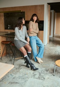 Deux femmes assises dans une cuisine moderne. L'une porte un pull zippé gris et une jupe à carreaux ; l'autre un haut en tricot beige et un jean bleu.