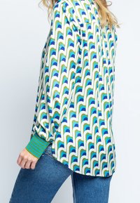Blusa colorida con un patrón geométrico de ondas en azul, verde y blanco, con mangas sueltas y un puño verde a rayas.