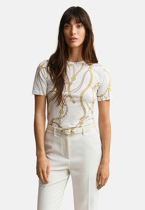 Vrouw met lang bruin haar, gekleed in een witte broek en een wit shirt met korte mouwen met een gouden kettingenprint, staand tegen een effen achtergrond.