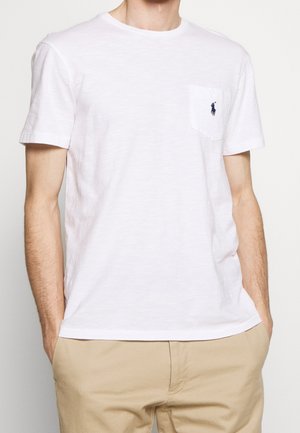 Homme portant un t-shirt blanc uni à manches courtes avec un petit logo brodé sur la poche poitrine, associé à un pantalon beige.