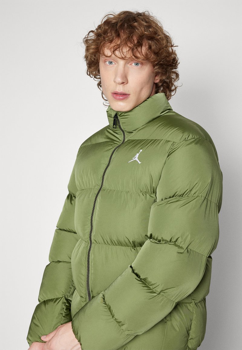 Giacca a vento verde con colletto alto, dotata di zip frontale e un accento con logo bianco. Il tessuto ha una texture liscia e imbottita.