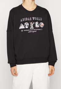 Svart sweatshirt med rund halsringning och långa ärmar. Framsidan har en grafisk design med färgglada märken och text, inklusive "ADIDAS WORLD."