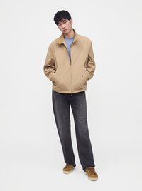 Homme portant une veste beige zippée, une chemise bleue, un jean noir et des baskets marron, debout les mains dans les poches sur fond blanc.