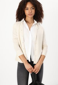 Beige Strickjacke mit langen Ärmeln und Knopfverschluss, kombiniert mit einem weißen Hemd und dunklen Jeans. Hält eine schwarze Tasche.