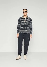 Fleece-pullover med geometriske mønstre i marine og krem, kombinert med svarte joggebukser. Har halv-zip krage og en avslappet passform, med beige sko.