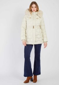 Donzen crème winterjas met een capuchon van nepbont, een taille met ceintuur en zakken met rits. Gecombineerd met marineblauwe corduroy broek met uitlopende pijpen en bruine veterhakken.