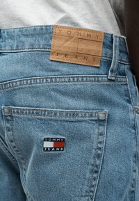 Κοντινή λήψη ενός ανοιχτόχρωμου μπλε τζιν με καφέ δερμάτινη ταμπέλα και το λογότυπο Tommy Jeans στην πίσω τσέπη.