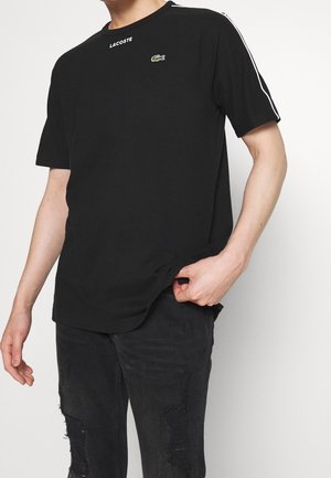 Camiseta negra de manga corta, con escote redondo y un logo de cocodrilo verde de Lacoste en el pecho. Cuenta con rayas blancas en los costados.